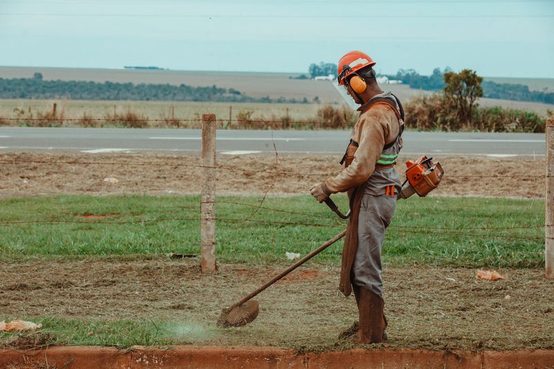 Jardineiro com roçadeira e EPI trabalhando em área verde extensa