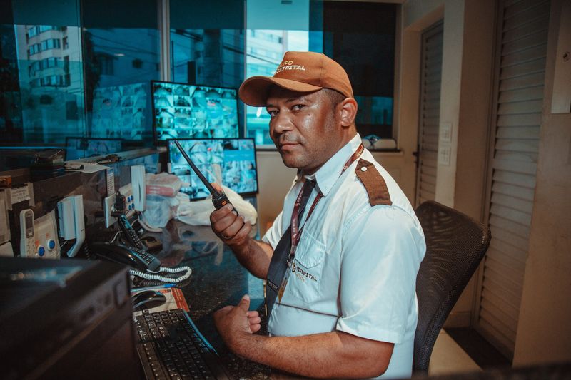 Porteiro do Grupo Distrital em guarita, operando o controle de acesso com rádio e monitores