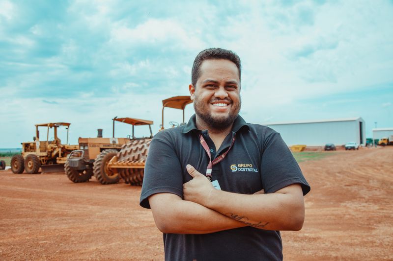 Colaborador do Grupo Distrital sorrindo em frente de obra com máquinas ao fundo